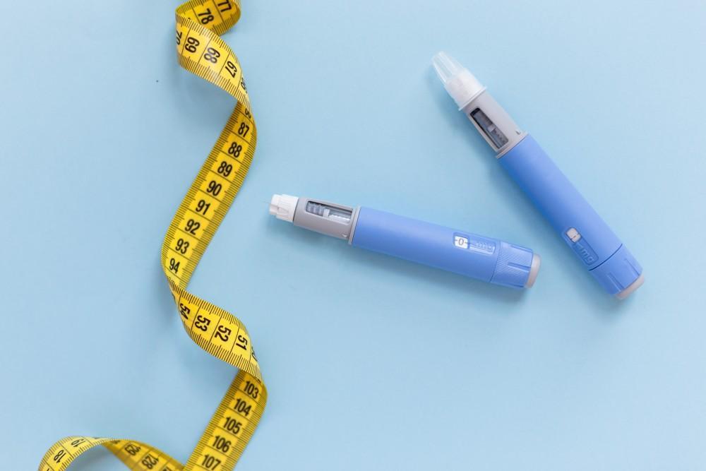 Weight loss injection pens with yellow measuring tape on blue background – representing medical weight management and hormone therapy tools.