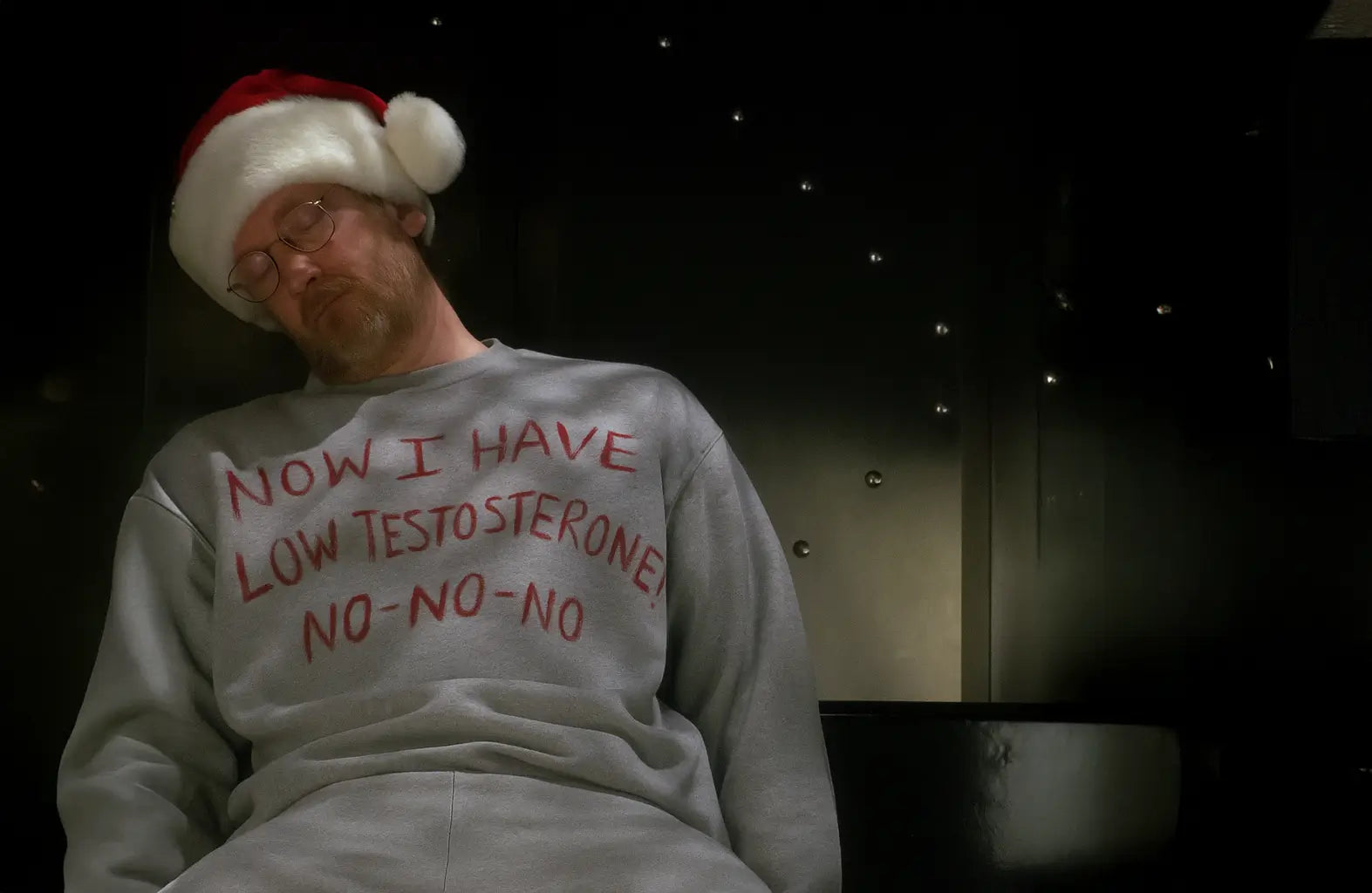 Middle-aged man wearing a Santa hat and grey jumper reading ‘Now I Have Low Testosterone, No-No-No,’ asleep in a dimly lit lift, symbolising low energy and low testosterone symptoms in men over 40.